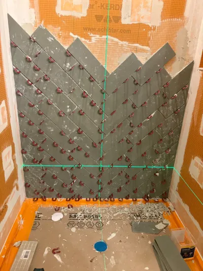 Shower wall mid-installation with grey chevron tile pattern and laser level lines guiding the layout.