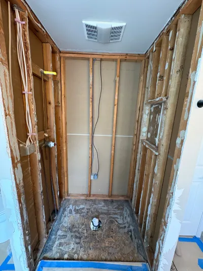 A gutted bathroom alcove with exposed wood studs, rough-in plumbing, and a plywood subfloor mid-renovation.