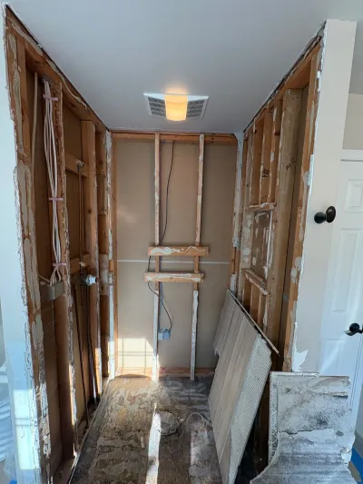 Bathroom under renovation with exposed wall studs, rough-in plumbing and wiring, and a lit exhaust fan.