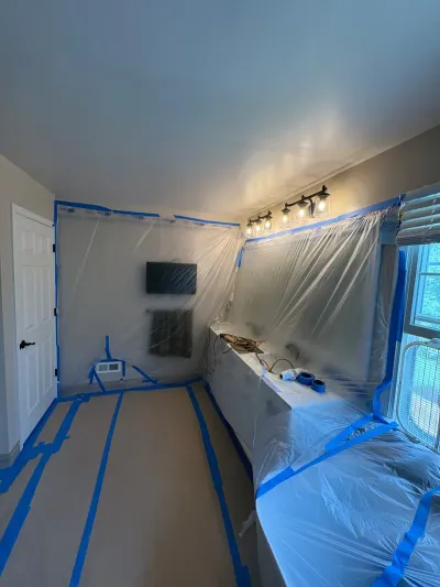 Bedroom prepped for painting with plastic sheeting and blue tape covering walls, window, and furniture.