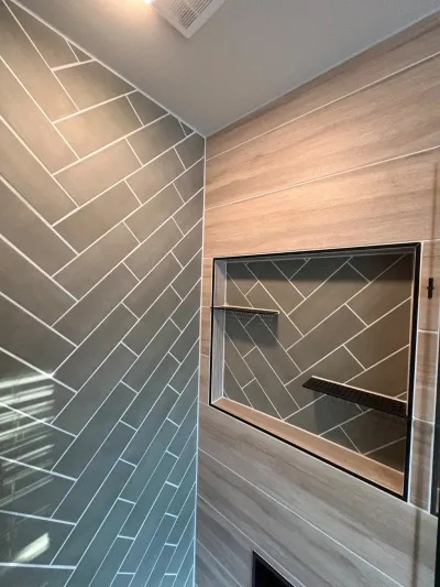 Walk-in shower corner with charcoal herringbone tile and wood-look tile wall framing a black-trimmed niche.
