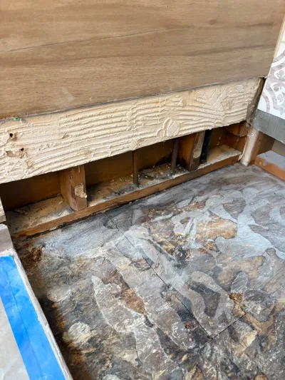 Close-up of exposed wall studs with spray foam insulation above a stained, worn subfloor during renovation.