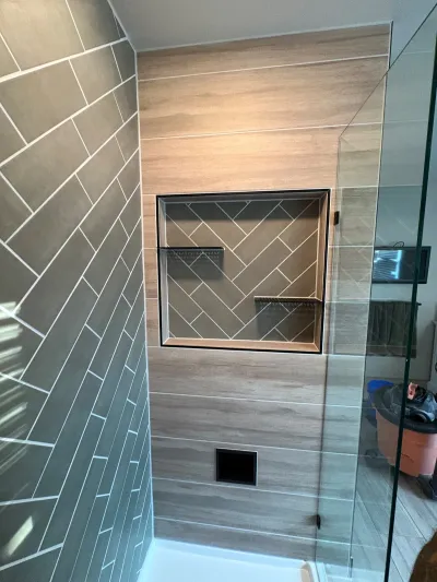 Finished walk-in shower with wood-look tile, a herringbone accent wall, and a tiled niche with black shelves.